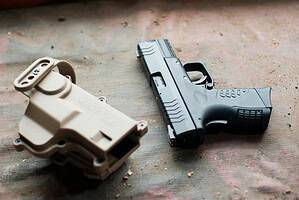 Close-up of a black handgun and beige holster on a textured surface, showcasing firearm details.