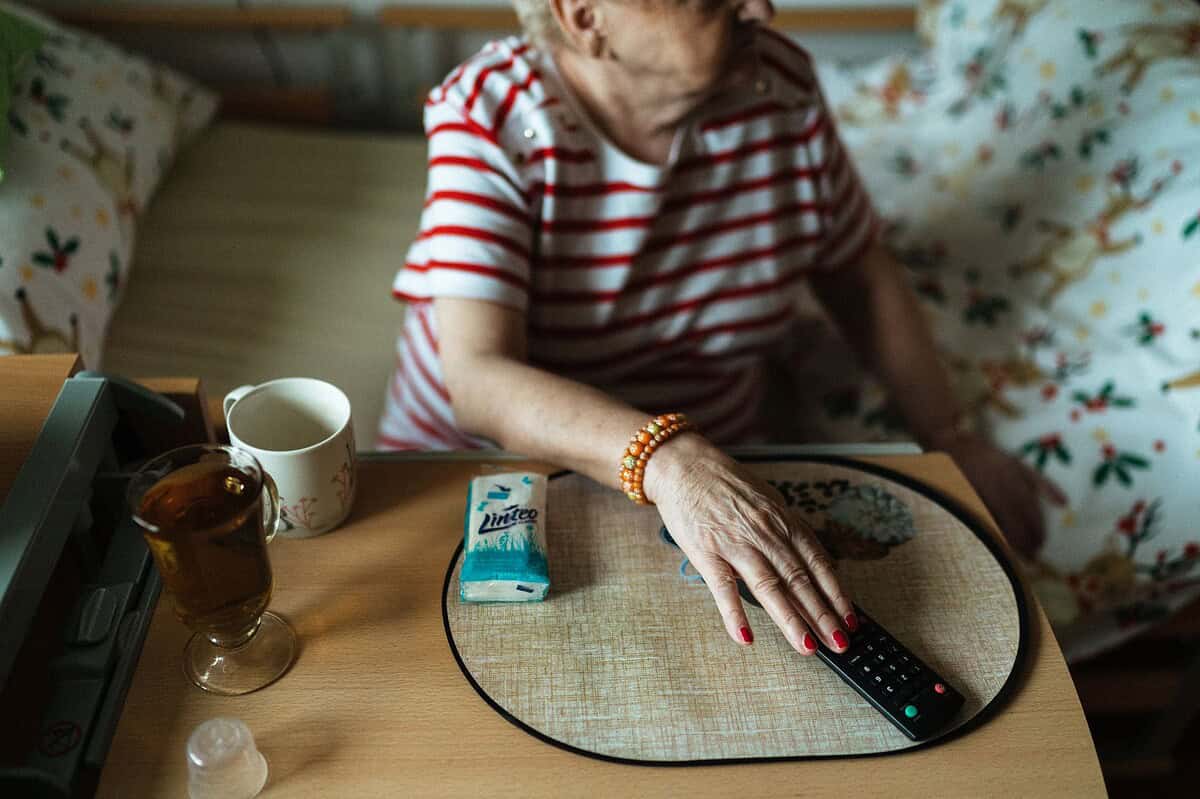 Senior woman in nursing home enjoying tea, TV remote in hand, in a cozy room.