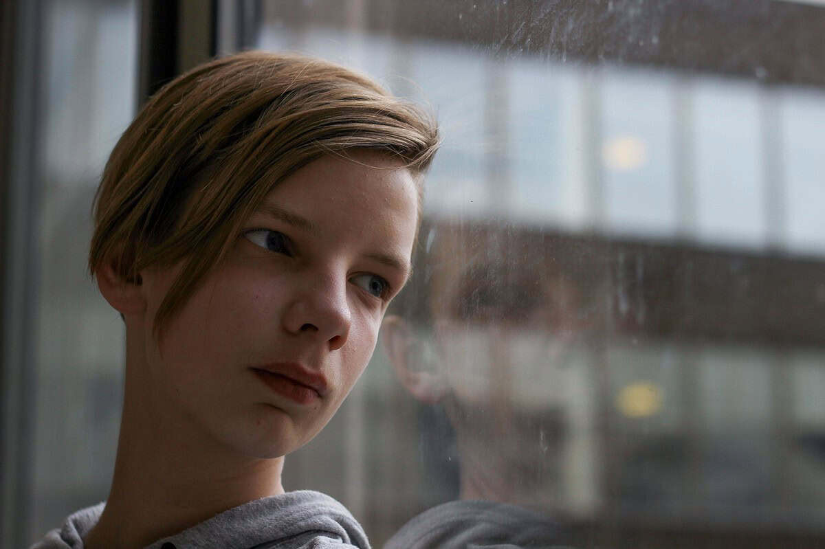 A thoughtful teenager gazing out a window, reflecting in the glass.